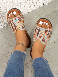 Open-toe flat sandals with tan patterned straps and brown trim, worn with jeans
